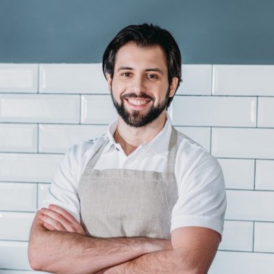 Portrait of smiling shop assistant in apron with arms crossed standing at counter in supermarket e1674721586755.jpg